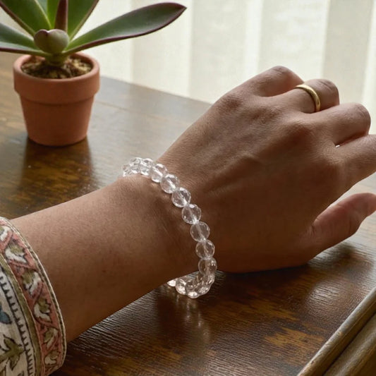 Hand wearing a clear quartz bracelet, diamond cut stones on a wooden surface with a potted plant in the background
